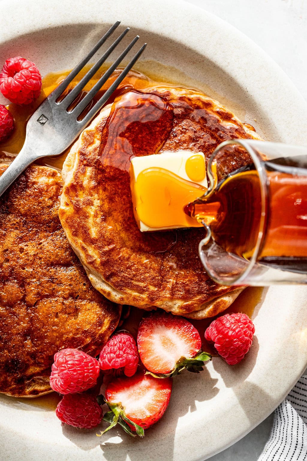 Two pancakes topped with butter and syrup, garnished with fresh raspberries and halved strawberries, with a fork on the plate.