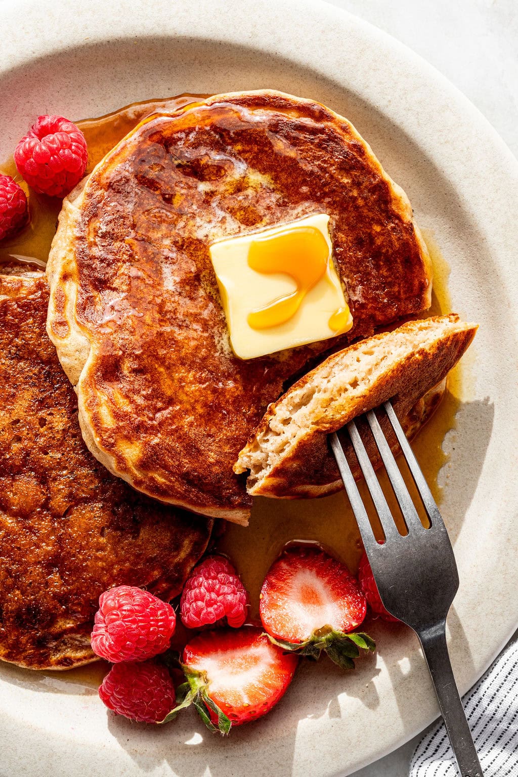Two pancakes topped with a pat of butter and syrup, served with raspberries and strawberries on a beige plate. A fork holds a cut piece of pancake.