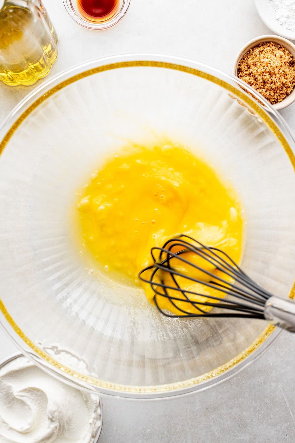 A metal whisk mixing beaten eggs in a large glass bowl, surrounded by ingredients including brown sugar, vanilla extract, and flour.