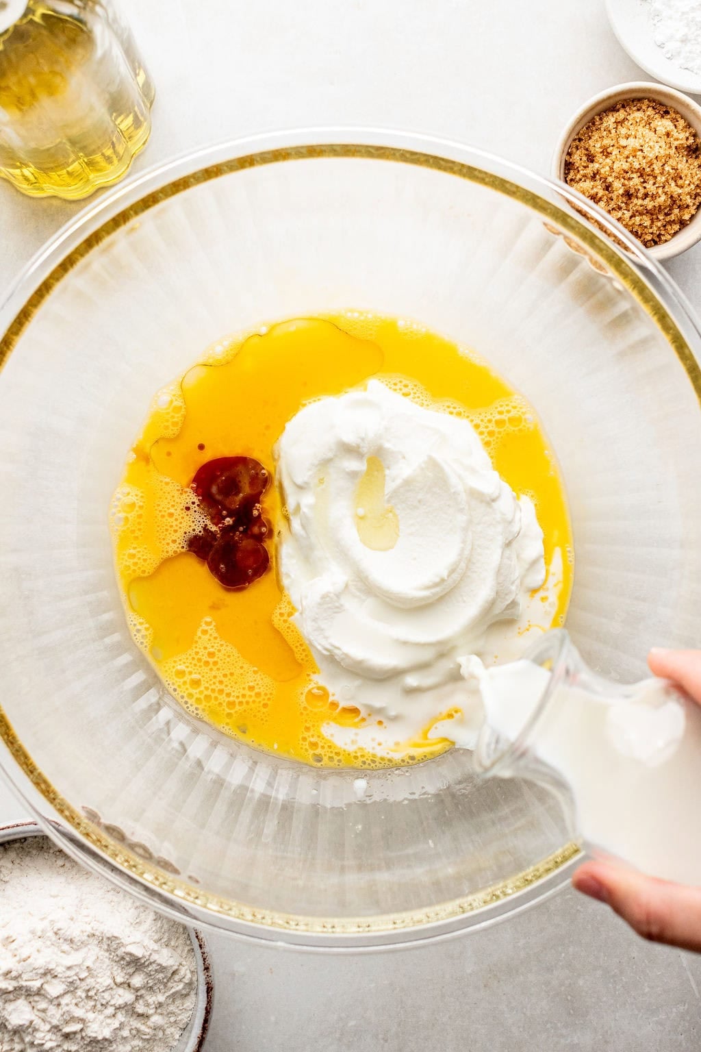A large bowl with eggs, Greek yogurt, and vanilla being mixed while milk is poured in; surrounding are bowls of flour, brown sugar, and oil.