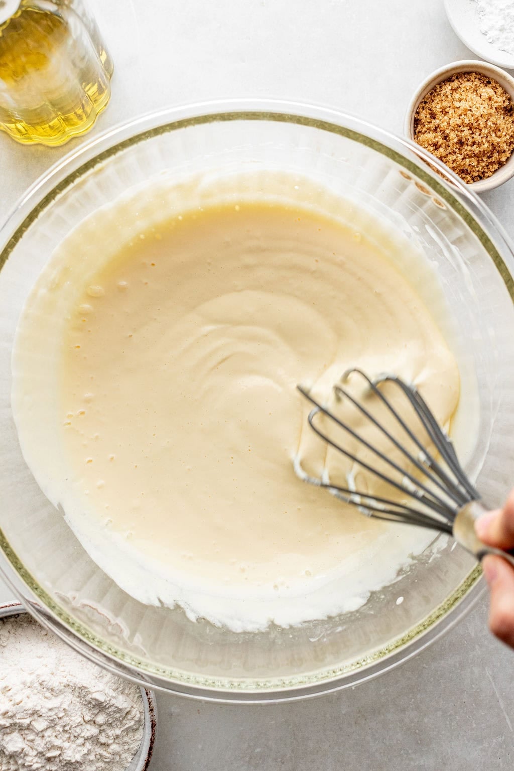A hand uses a whisk to mix a smooth batter in a large glass bowl, surrounded by flour, brown sugar, and a jar of oil on a light surface.
