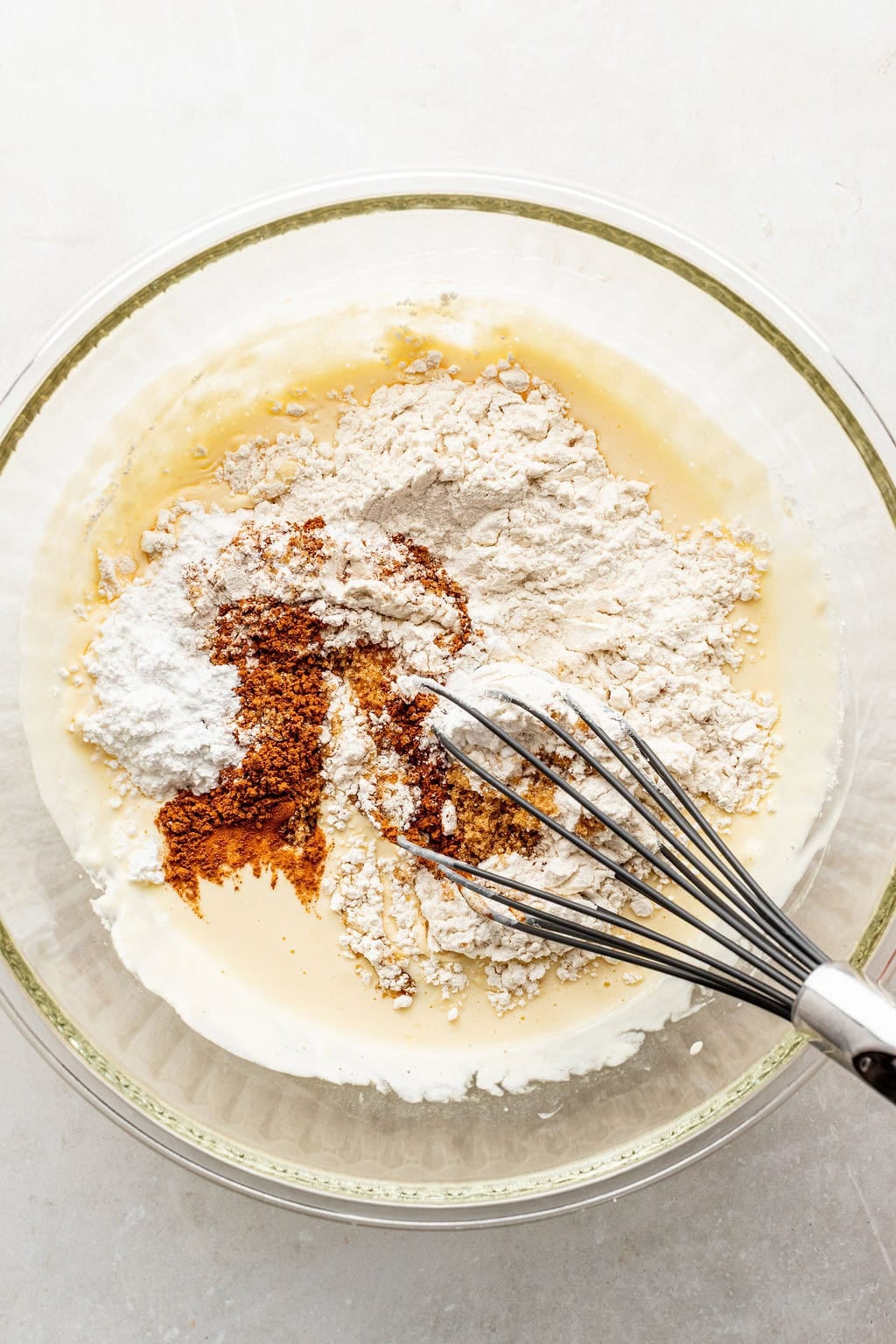 A glass bowl with flour, baking powder, spices, and wet ingredients being mixed with a metal whisk on a light counter.