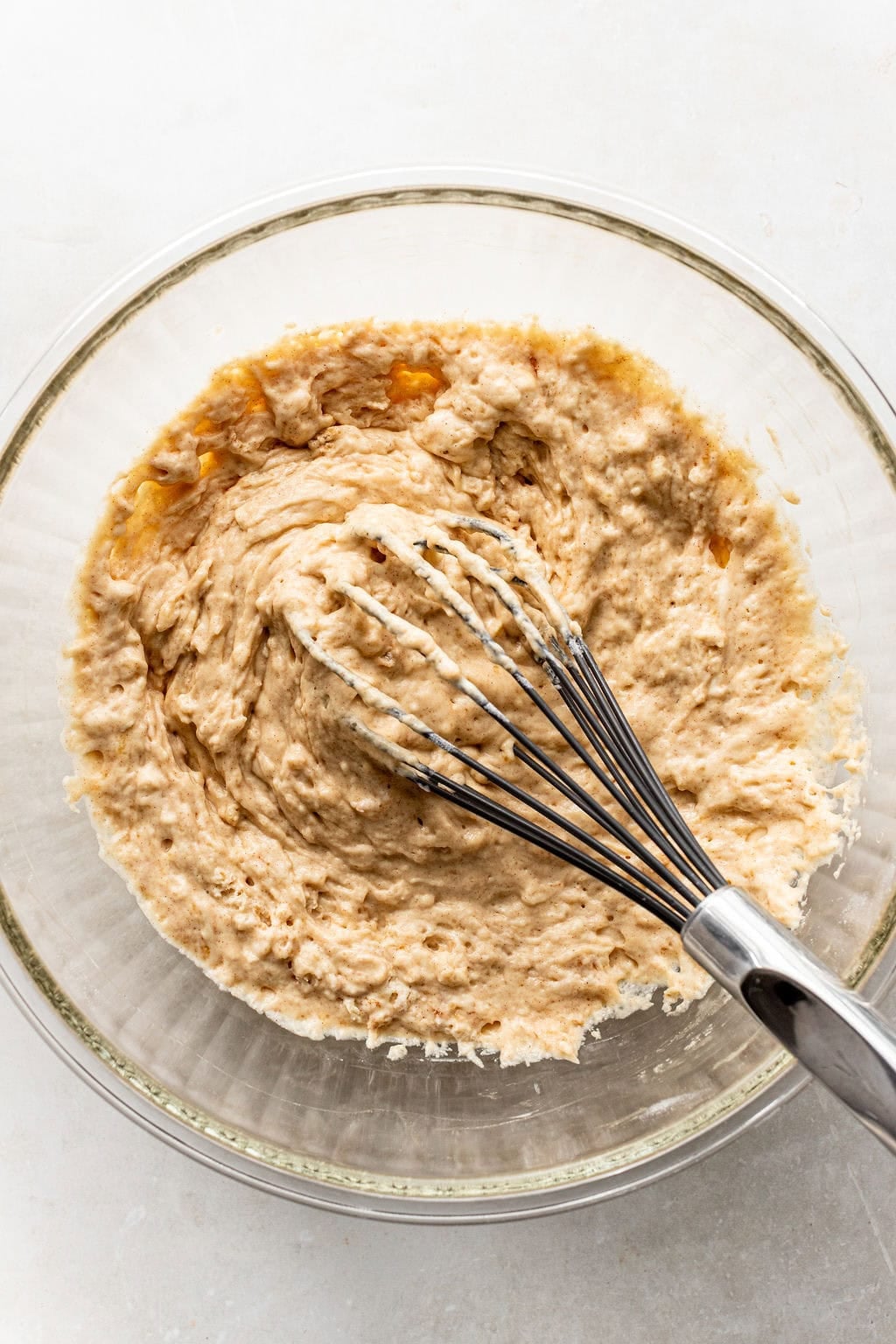A metal whisk mixing light brown batter in a clear glass bowl on a light surface.