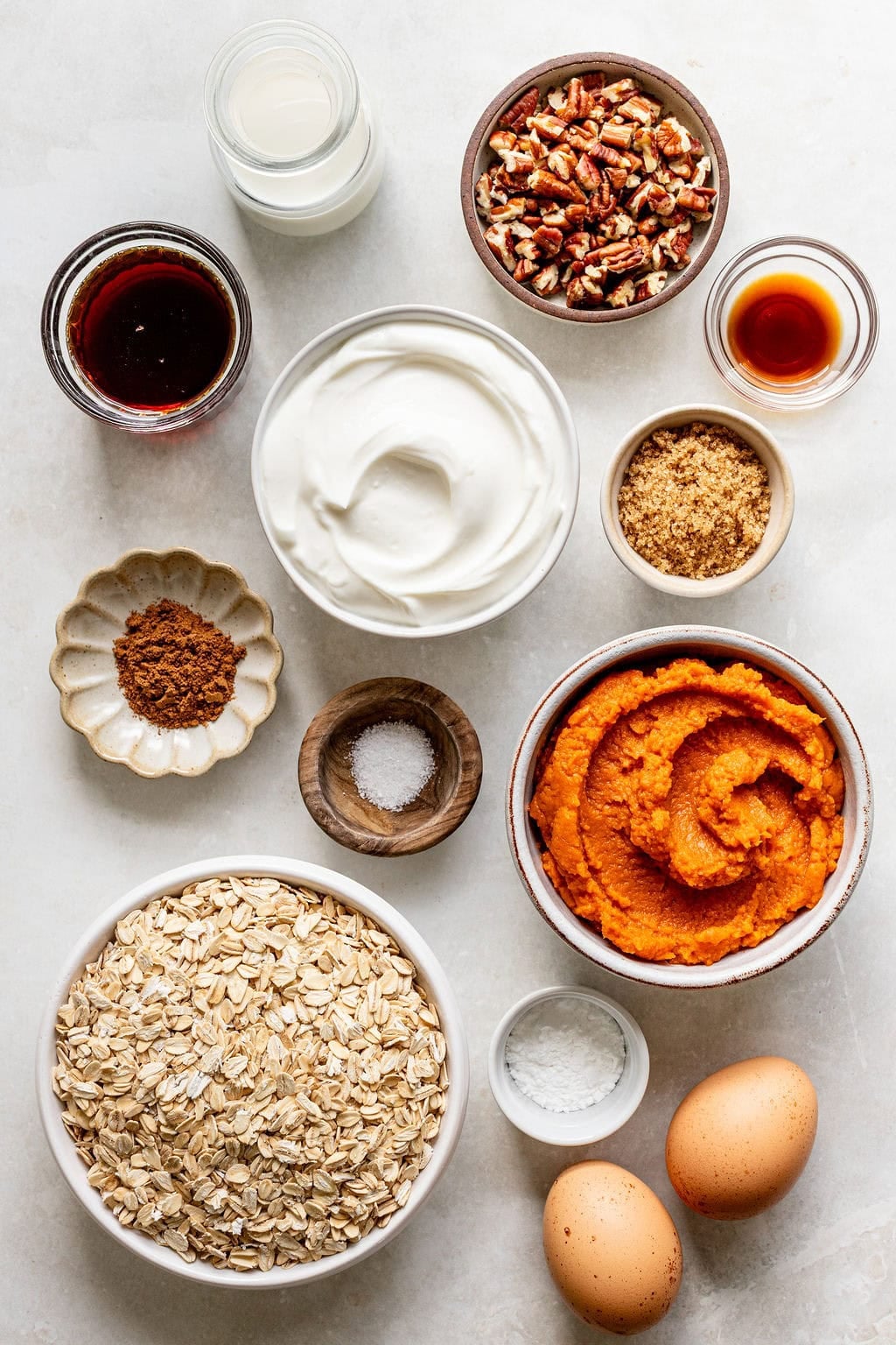 Various baking ingredients in bowls on a countertop, including pumpkin puree, oats, eggs, Greek yogurt, pecans, spices, brown sugar, vanilla extract, milk, and salt.
