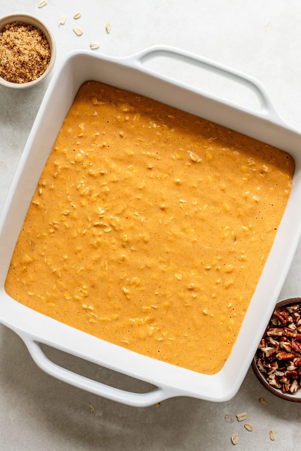 Square white baking dish filled with unbaked pumpkin oat mixture, surrounded by small bowls of brown sugar, oats, and chopped pecans on a light surface.
