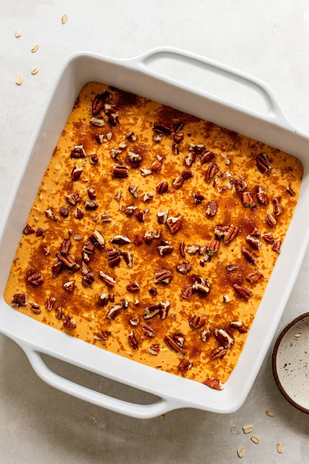 Square white baking dish filled with a golden orange baked mixture, topped with chopped pecans, on a light surface.