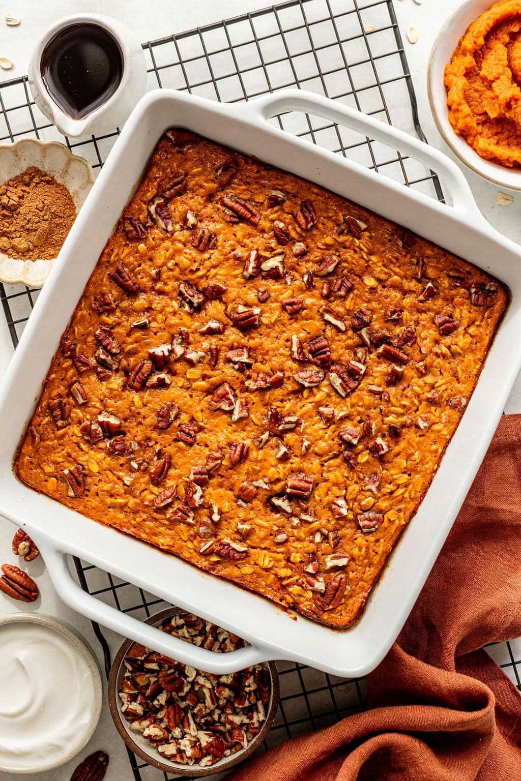 A square baking dish with baked oatmeal topped with pecans sits on a cooling rack, surrounded by small bowls of syrup, pumpkin puree, whipped cream, spices, and chopped pecans.