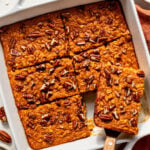 Baked pumpkin oat bars with pecans cut into squares in a white baking dish, one piece being lifted with a spatula.