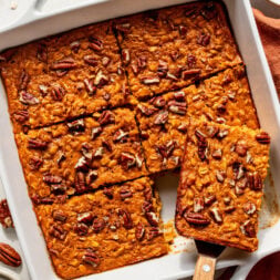 Baked pumpkin oat bars with pecans cut into squares in a white baking dish, one piece being lifted with a spatula.