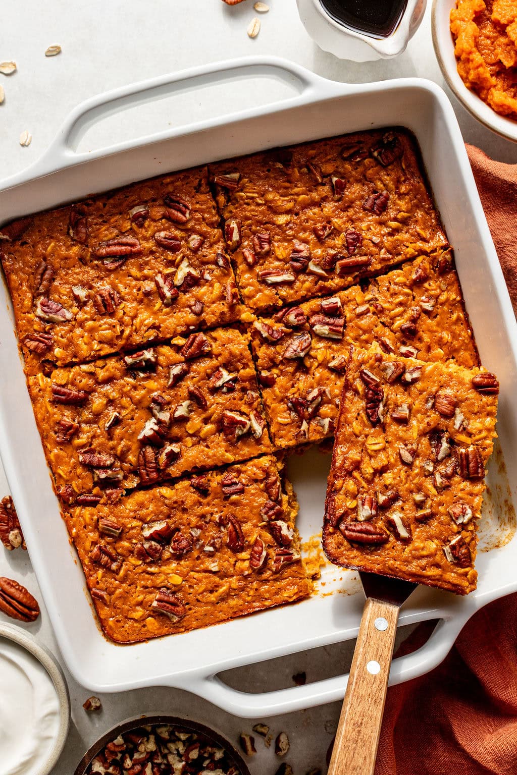 Baked pumpkin oat bars with pecans cut into squares in a white baking dish, one piece being lifted with a spatula.