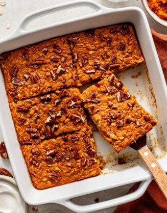 Baked pumpkin oat bars with pecans in a white pan, one bar being lifted with a spatula. Nearby are small bowls of whipped cream, chopped pecans, and syrup.