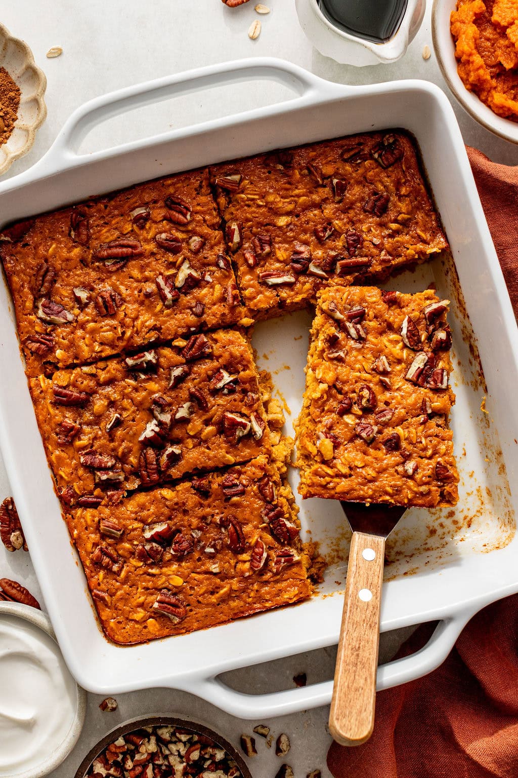 Baked pumpkin oat bars cut into squares in a white dish, with a spatula lifting one piece; chopped pecans sprinkled on top.