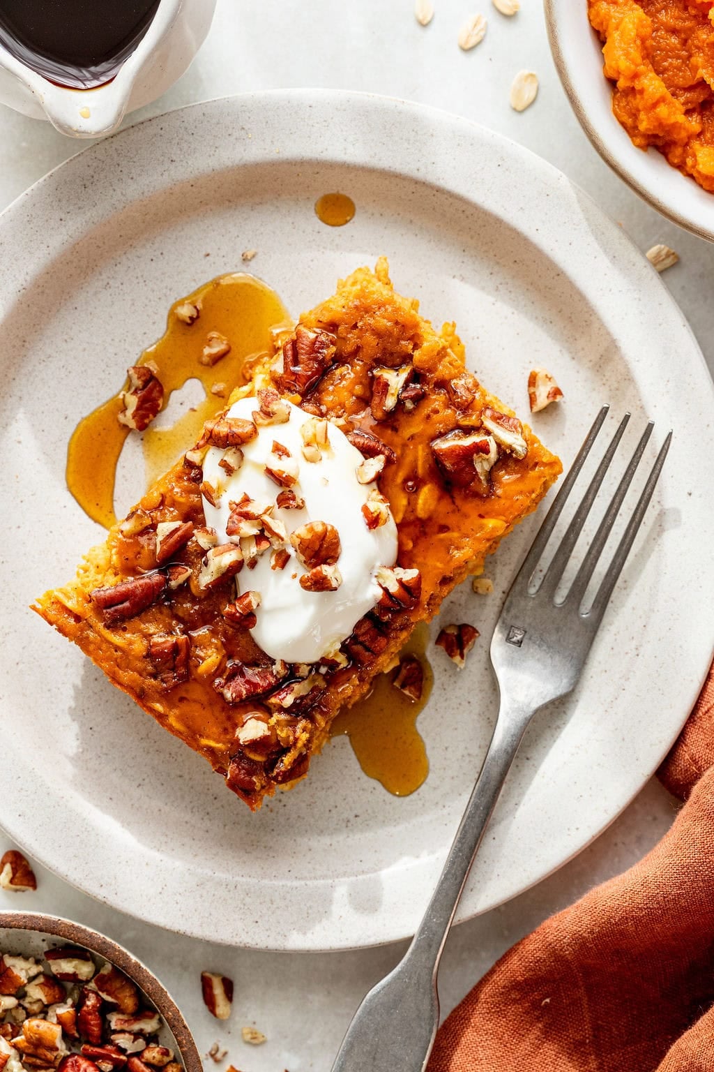 A slice of pumpkin baked oatmeal topped with whipped cream, chopped pecans, and maple syrup sits on a white plate with a fork.