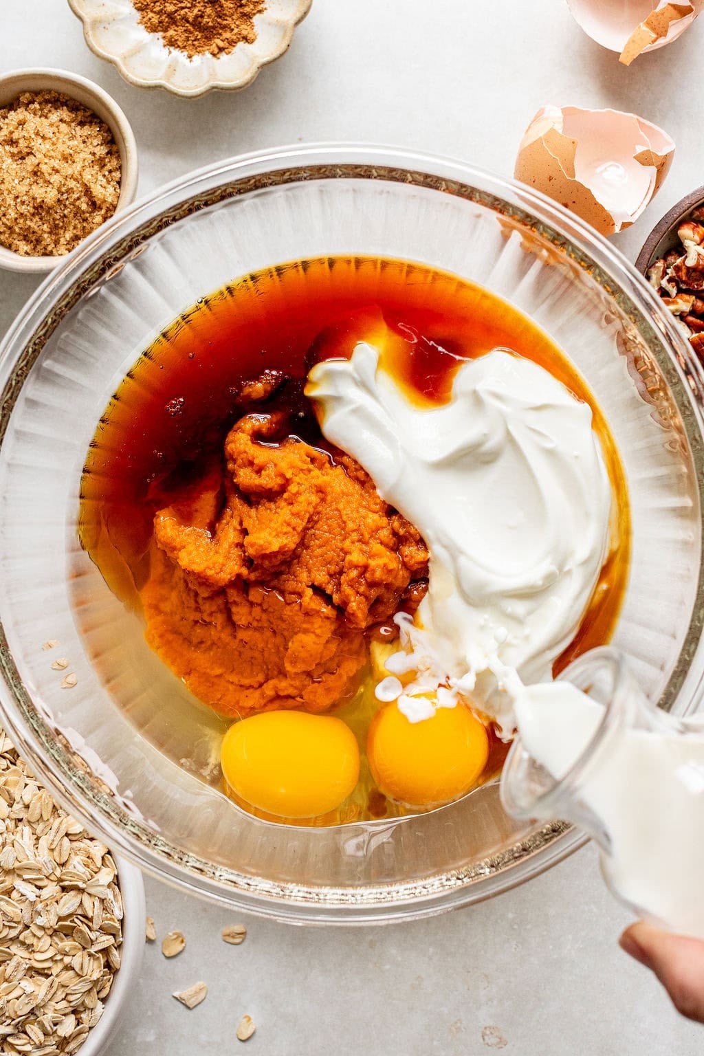 A glass bowl with eggs, pumpkin puree, sour cream, milk being poured, and brown liquid, surrounded by oats, brown sugar, pecans, cinnamon, and eggshells.