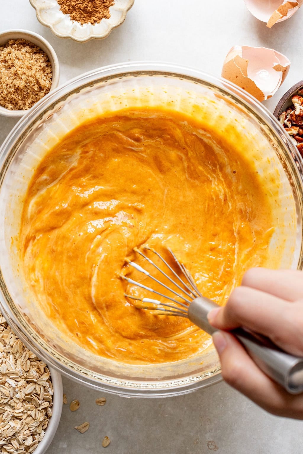 A hand uses a whisk to mix a bowl of orange batter. Surrounding the bowl are oats, brown sugar, chopped nuts, egg shells, and cinnamon.