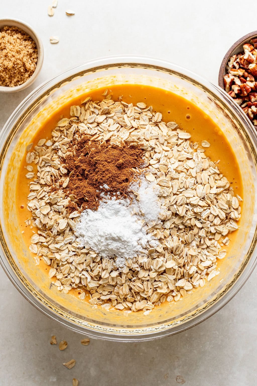 A glass bowl with oats, baking powder, cinnamon, and pumpkin mixture. Small bowls of brown sugar and chopped pecans are nearby on a light surface.