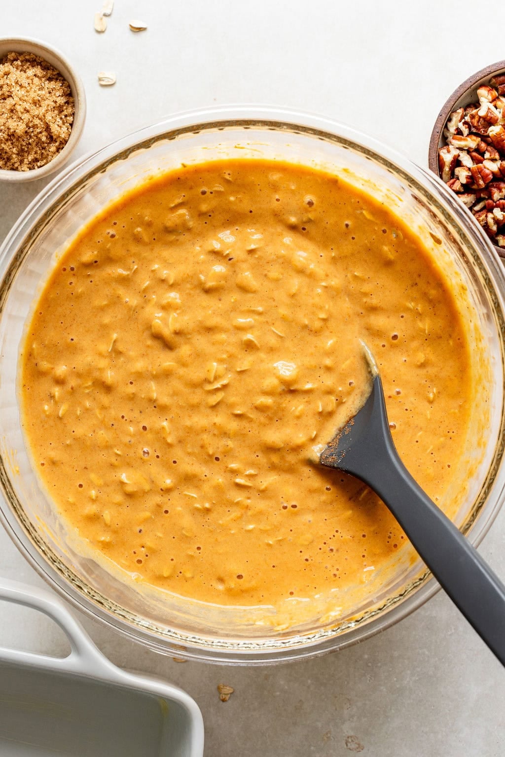 A glass bowl filled with pumpkin oat batter, a black spatula inside, surrounded by bowls of brown sugar, pecans, and an empty baking dish.