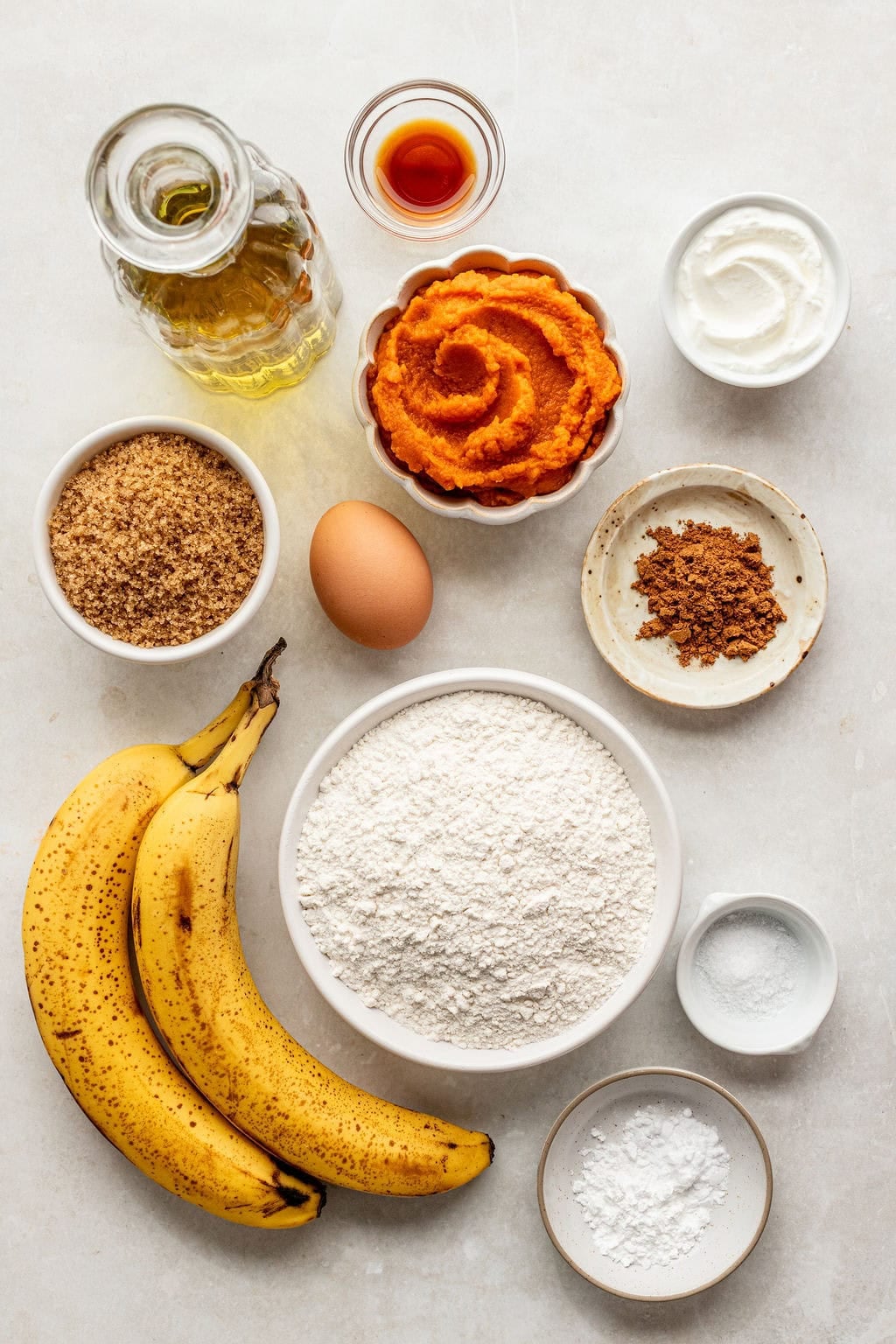 Bananas, flour, brown sugar, oil, pumpkin puree, an egg, vanilla, spices, yogurt, baking powder, baking soda, and salt arranged on a white surface.