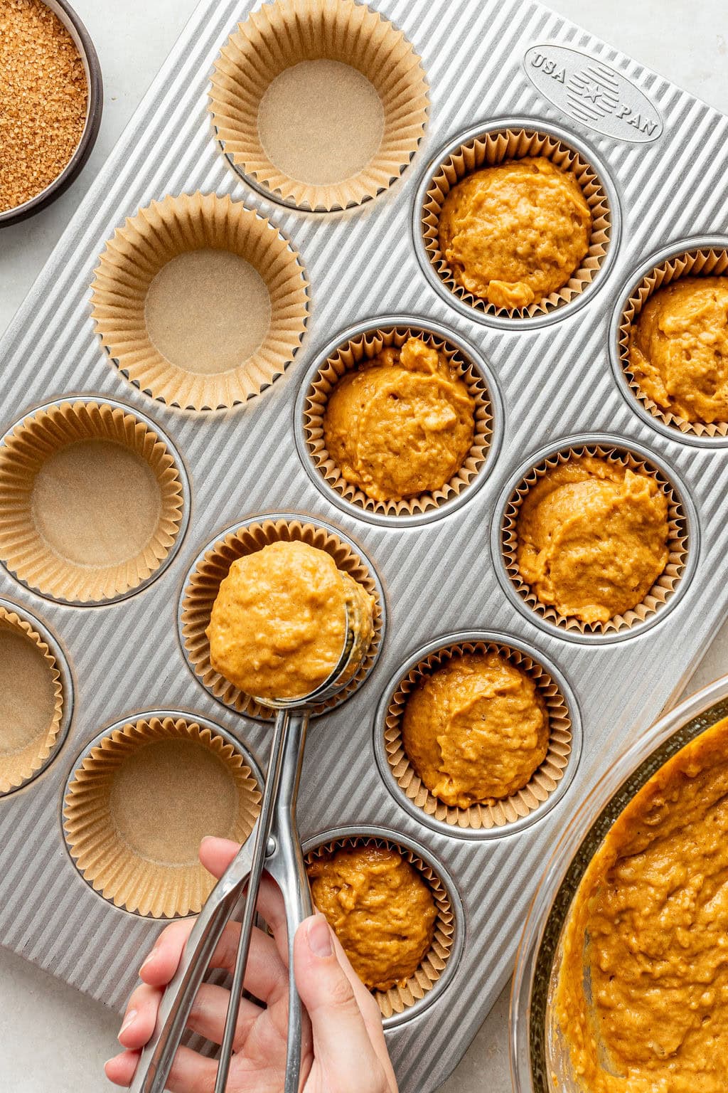 A hand scoops orange muffin batter into paper-lined muffin tins on a metal tray; a bowl of batter and empty liners are visible.