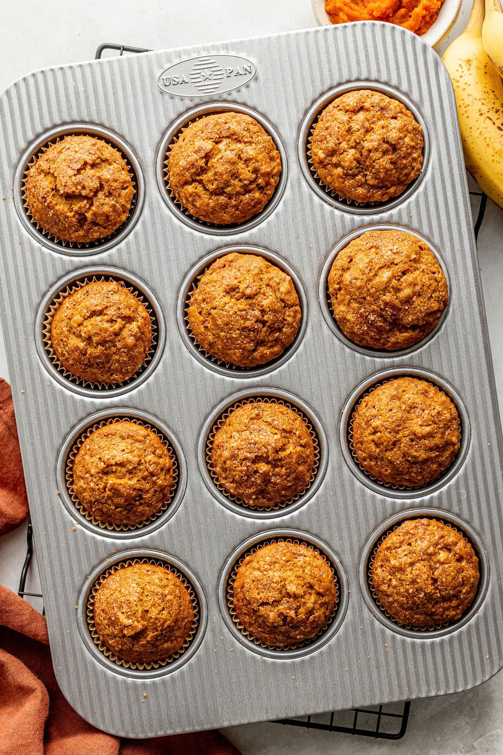A muffin tin filled with twelve baked muffins on a cooling rack, with part of a banana and an orange cloth visible at the edge.