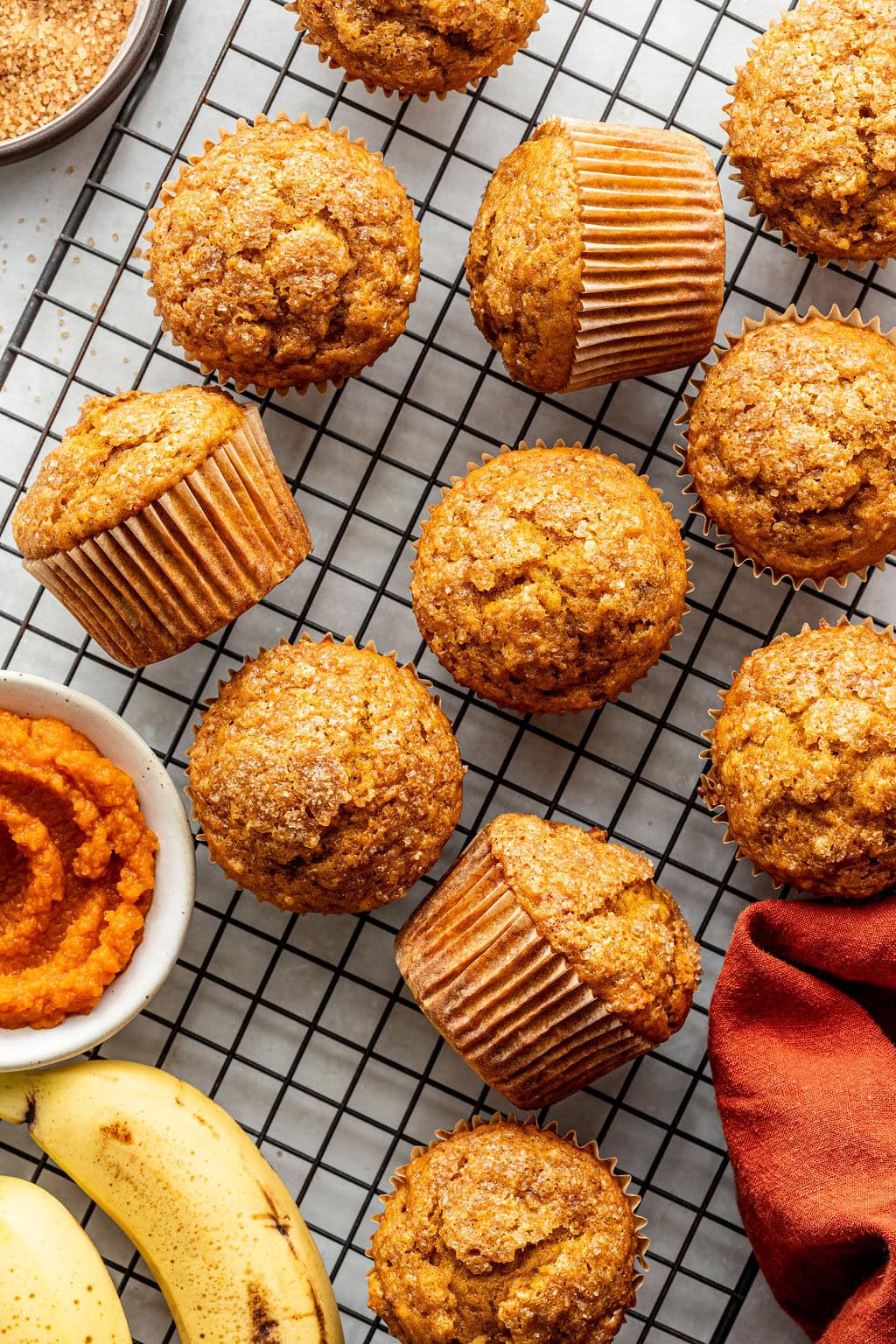 Banana muffins cooling on a wire rack, surrounded by a bowl of mashed banana, a banana, and a red cloth napkin.
