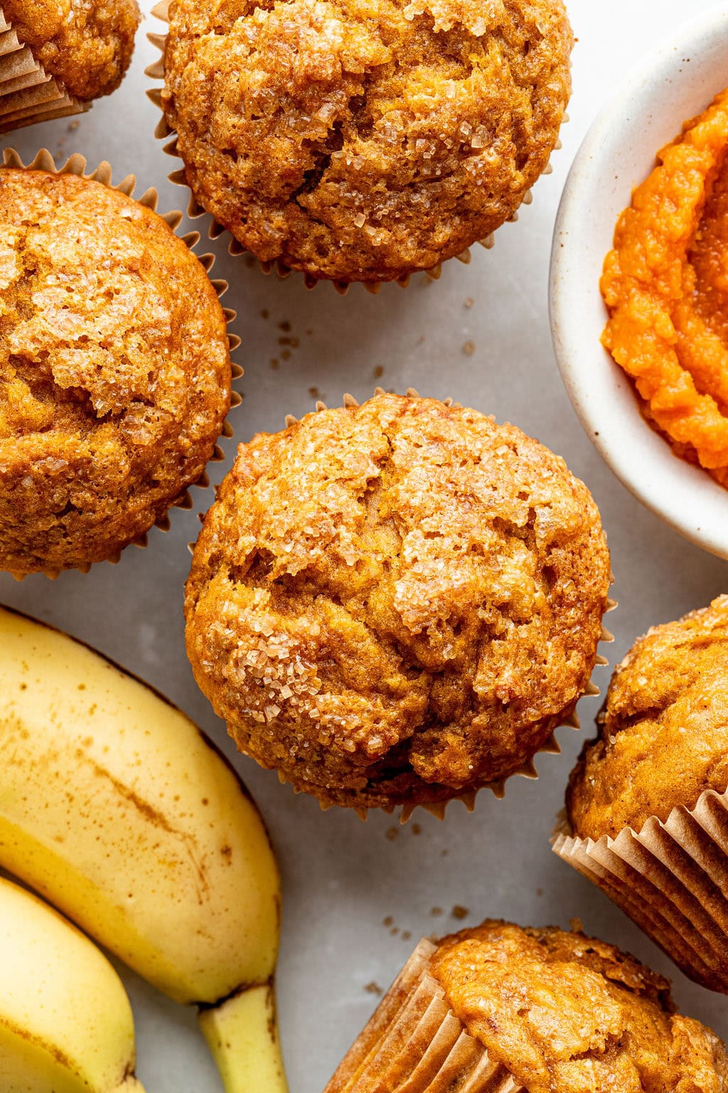 Banana muffins topped with sugar sit on a light surface next to whole bananas and a bowl of orange pumpkin puree.