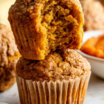 Two pumpkin muffins are stacked; the top muffin has a bite taken out, revealing a moist interior. Other muffins and ingredients are blurred in the background.