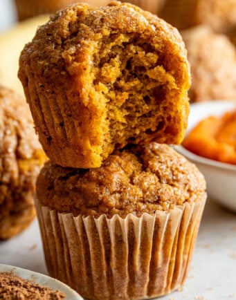 Two pumpkin muffins are stacked; the top muffin has a bite taken out, revealing a moist interior. Other muffins and ingredients are blurred in the background.