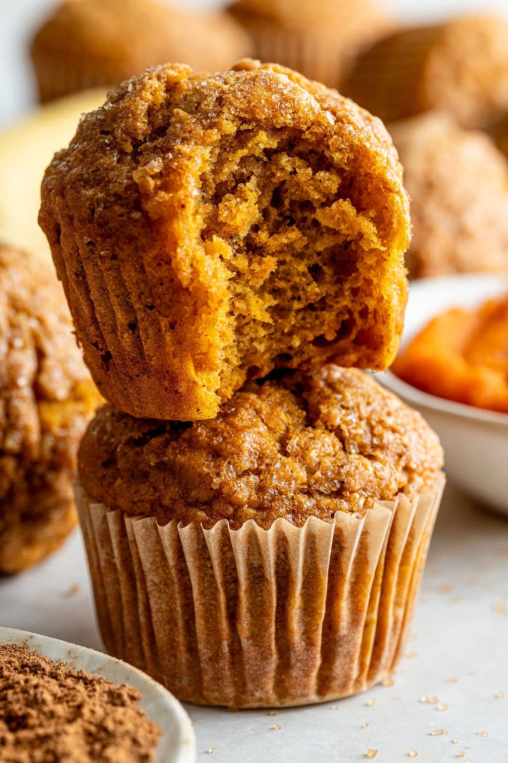 Two pumpkin muffins are stacked; the top muffin has a bite taken out, revealing a moist interior. Other muffins and ingredients are blurred in the background.