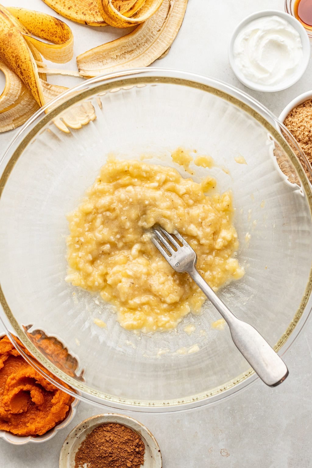 A fork mashes bananas in a glass bowl, surrounded by banana peels, yogurt, brown sugar, pumpkin puree, maple syrup, and spices on a light countertop.