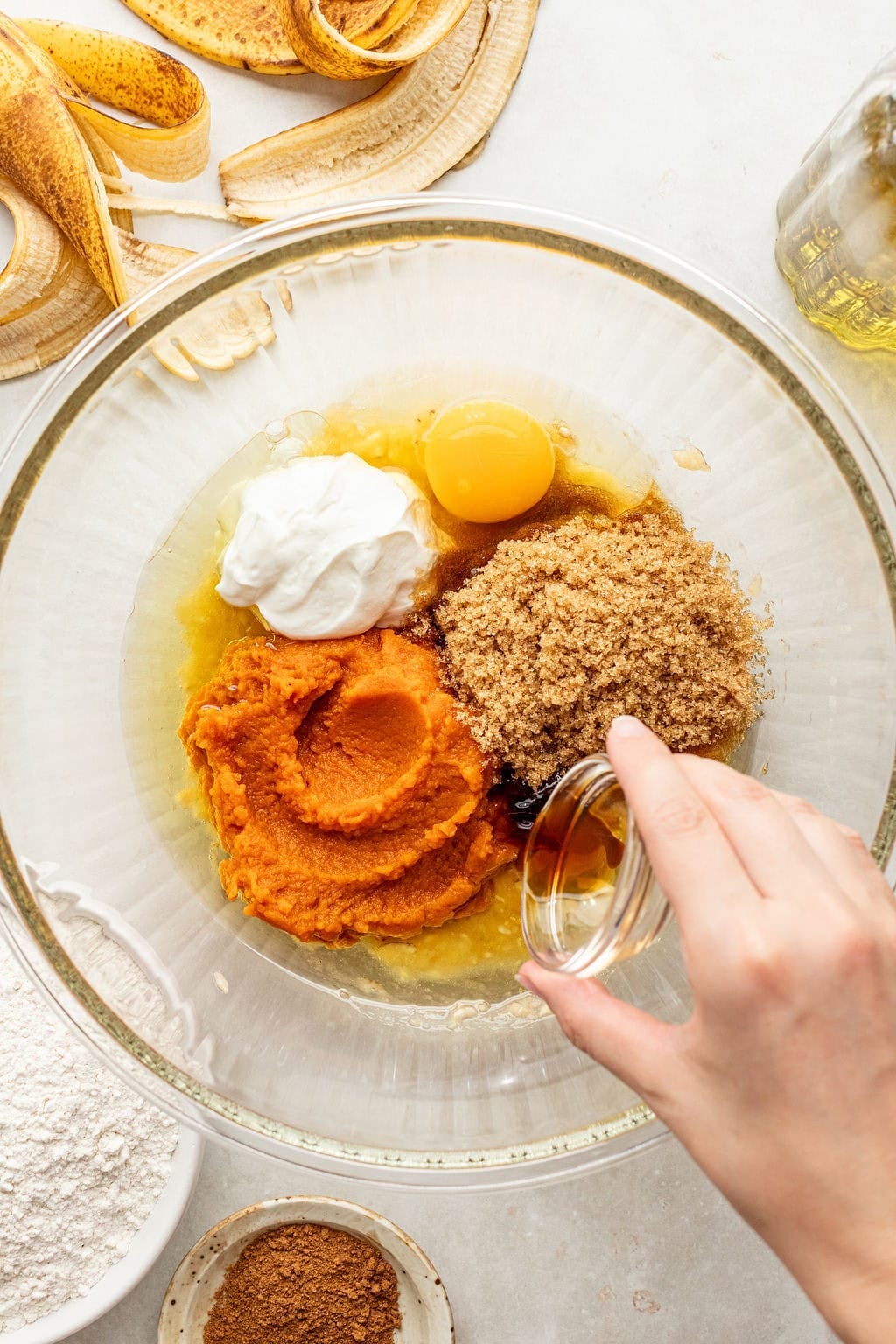 A hand adds liquid to a bowl containing pumpkin puree, brown sugar, egg, yogurt, and oil. Flour, spices, and banana peels are nearby on the counter.