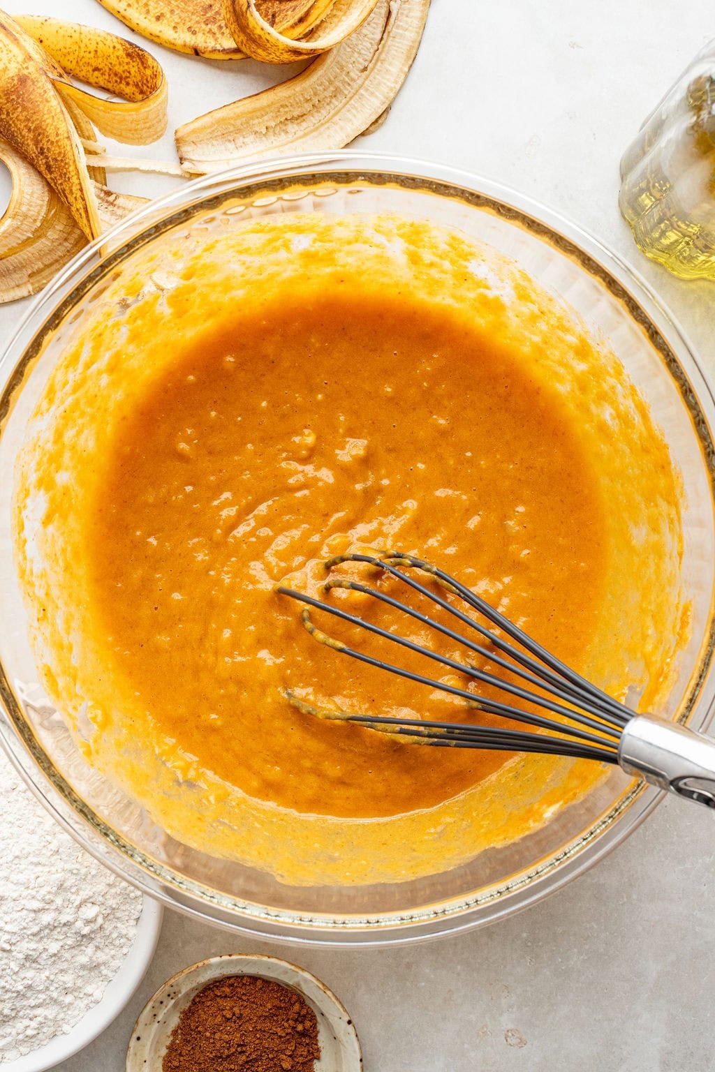 A glass bowl with banana bread batter and a whisk, surrounded by banana peels, flour, and a small bowl of spices on a white surface.