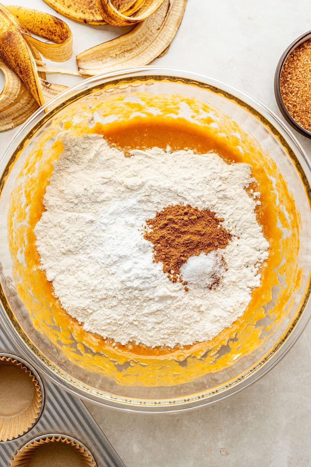 A glass bowl with flour, spices, and other baking ingredients on top of a wet mixture. Banana peels, brown sugar, and muffin liners are visible nearby.