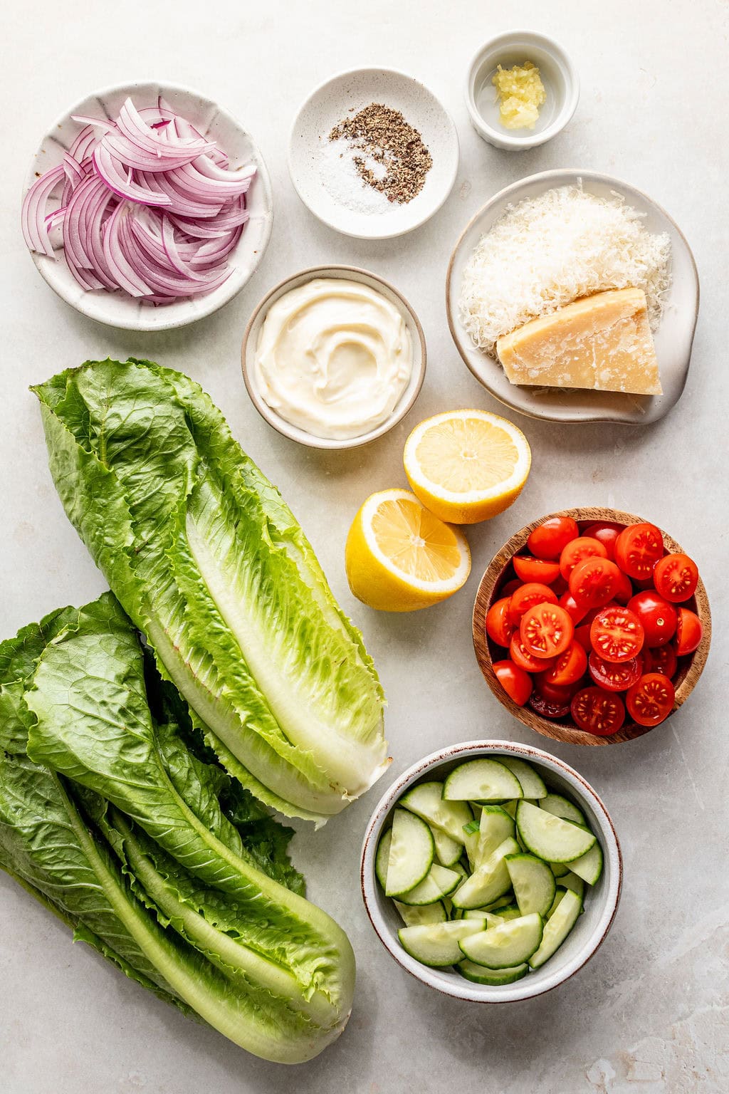 Ingredients for a salad arranged on a counter: romaine lettuce, cucumber, cherry tomatoes, lemon, mayonnaise, grated cheese, sliced red onion, minced garlic, salt, and pepper.