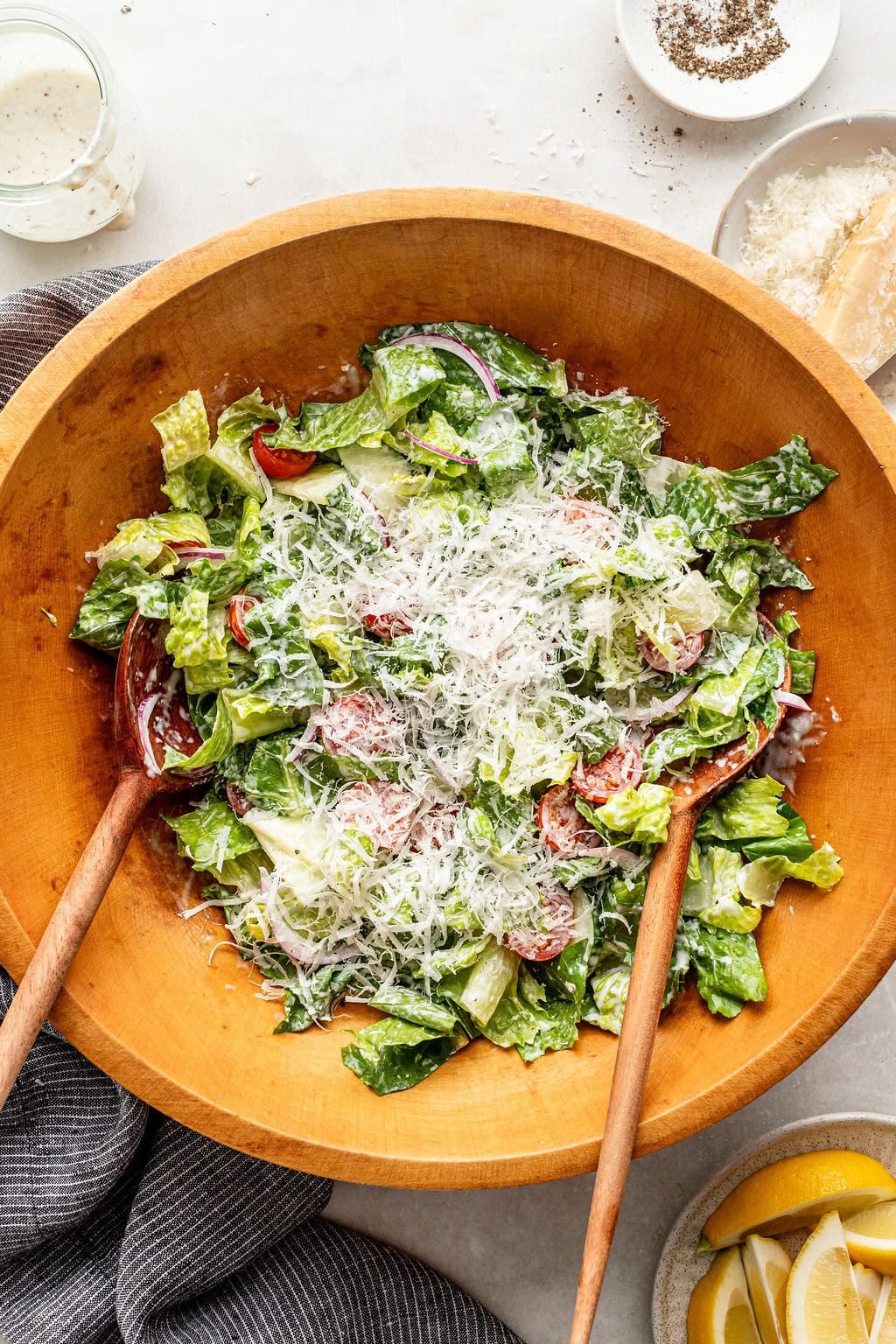 Large wooden bowl of salad with romaine lettuce, parmesan cheese, tomato slices, and two wooden serving utensils. Nearby are lemon wedges, pepper, grated cheese, and a jar of dressing.