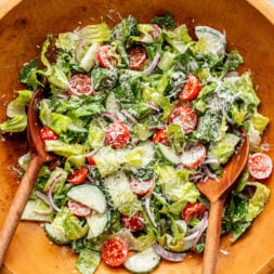Wooden bowl of salad with romaine lettuce, cherry tomatoes, sliced cucumber, red onion, shredded cheese, and creamy dressing; two wooden salad servers placed inside the bowl.