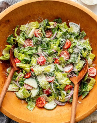 Wooden bowl of salad with romaine lettuce, cherry tomatoes, sliced cucumber, red onion, shredded cheese, and creamy dressing; two wooden salad servers placed inside the bowl.
