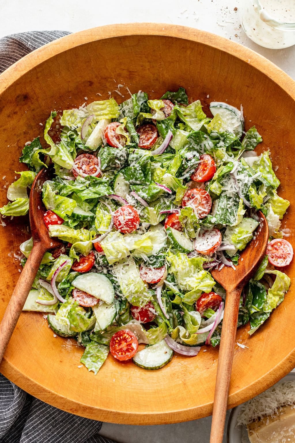 Wooden bowl of salad with romaine lettuce, cherry tomatoes, sliced cucumber, red onion, shredded cheese, and creamy dressing; two wooden salad servers placed inside the bowl.