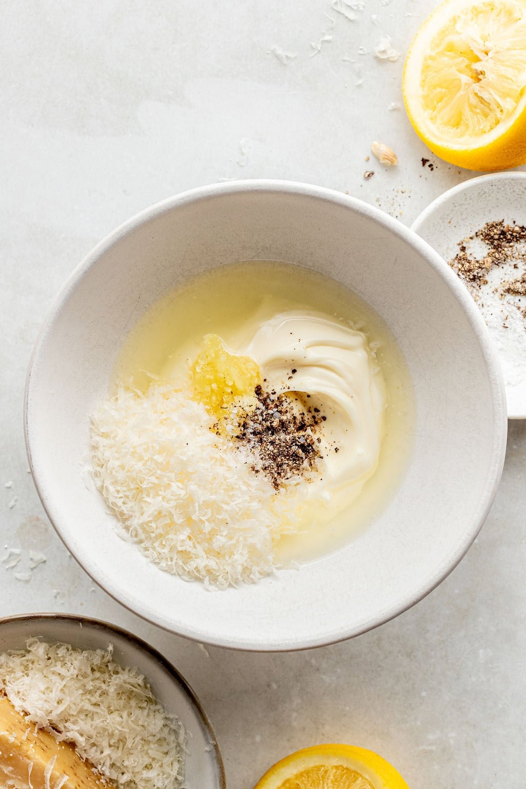 A white bowl contains mayonnaise, grated cheese, minced garlic, black pepper, and lemon juice. A halved lemon, extra cheese, and pepper are nearby on a light surface.