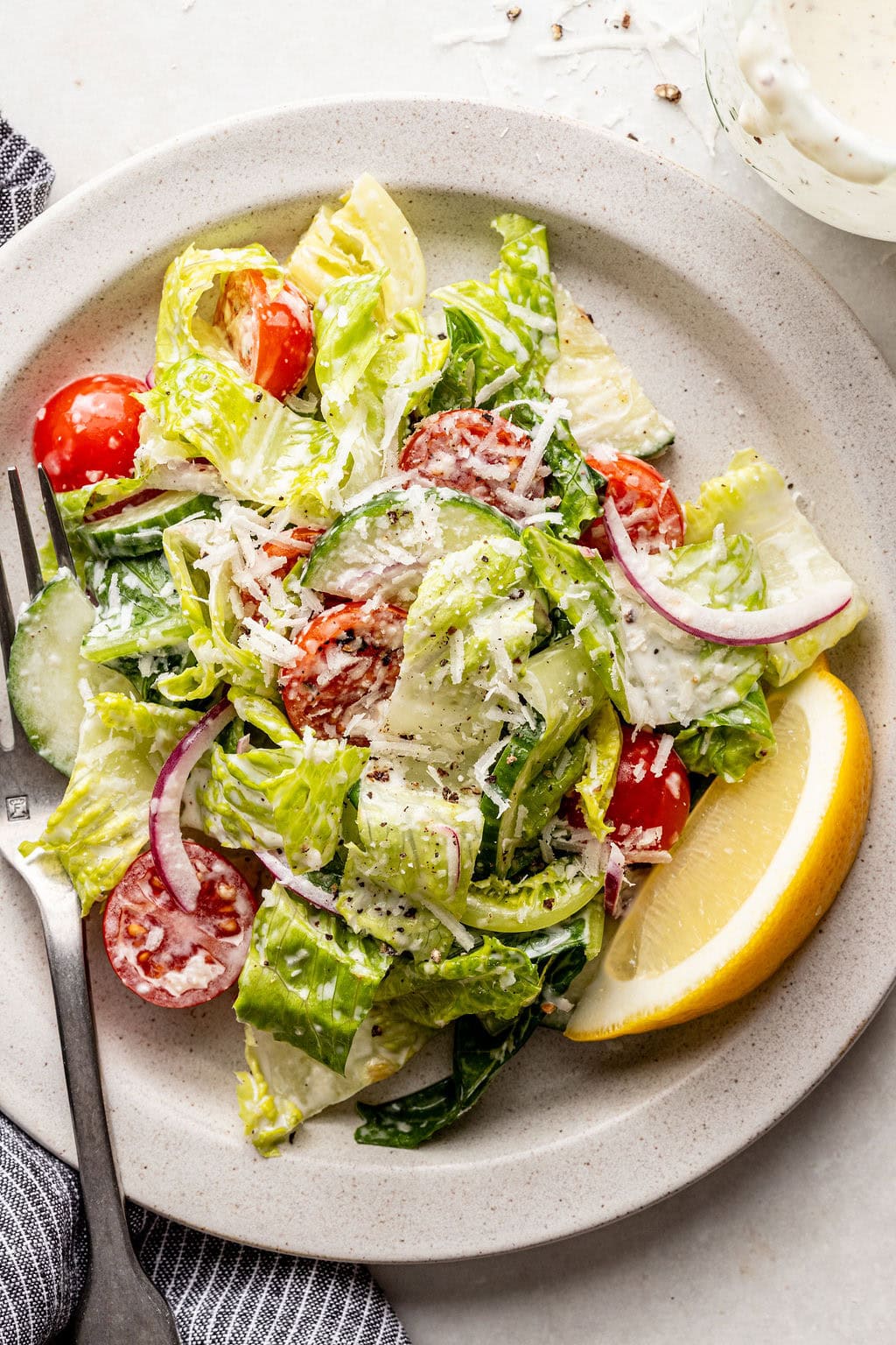 Plate of salad with lettuce, cherry tomatoes, cucumber, red onion, shredded cheese, a lemon wedge, and a fork, with creamy dressing on the side.