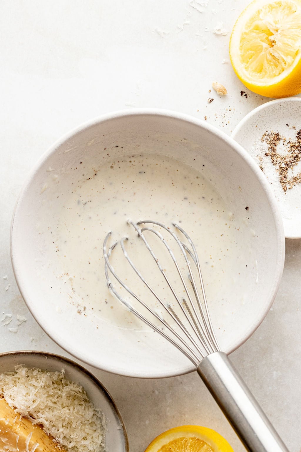 A metal whisk rests in a white bowl containing a creamy mixture; grated cheese, black pepper, and lemon halves are nearby on a light surface.