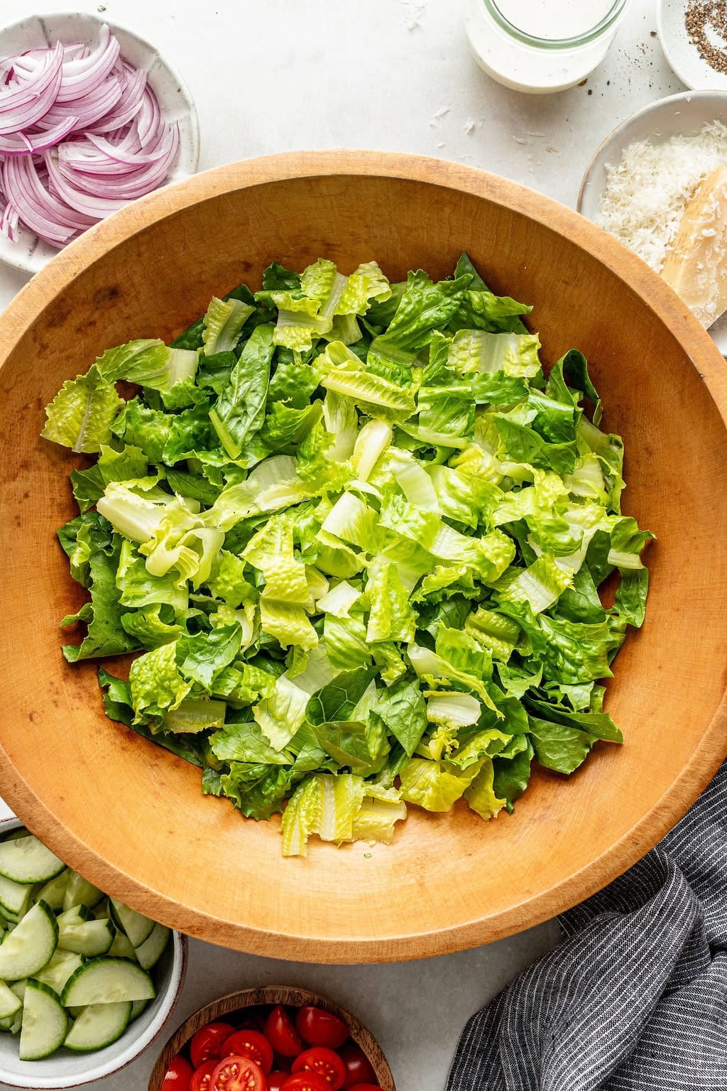 Wooden bowl filled with chopped romaine lettuce, surrounded by sliced red onion, cucumber, cherry tomatoes, grated cheese, and a small jar of salad dressing on a white surface.