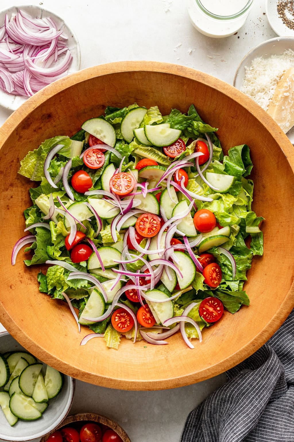 A wooden bowl filled with salad containing lettuce, cherry tomatoes, cucumber slices, and red onion, surrounded by small bowls of salad ingredients.