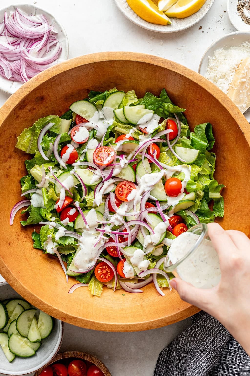 A hand pours dressing over a wooden bowl filled with salad containing lettuce, cucumber, cherry tomatoes, red onion, and sliced olives.