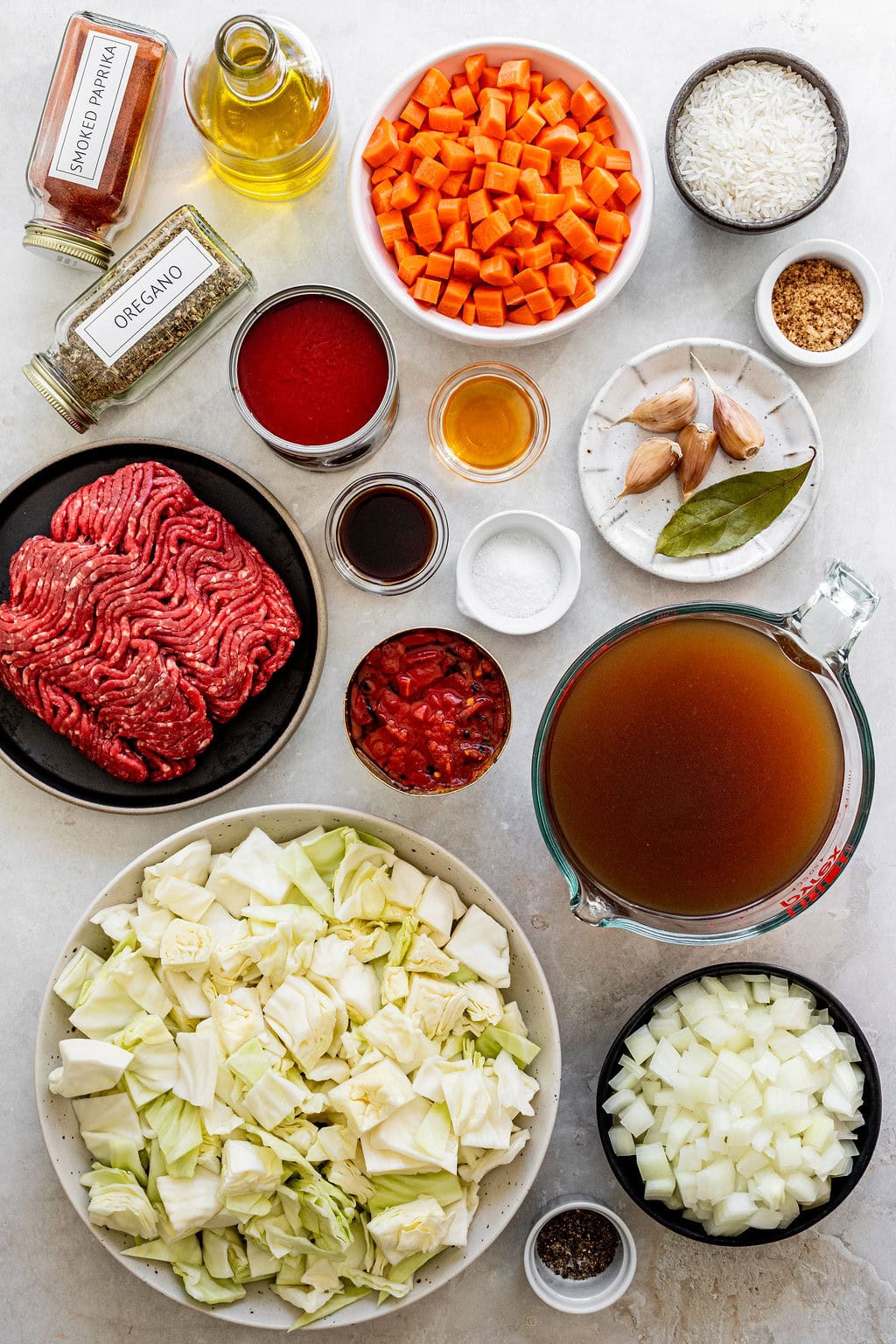 Top-down view of chopped vegetables, raw ground beef, spices, broth, rice, canned tomato, and liquid ingredients arranged in bowls and jars on a white surface.