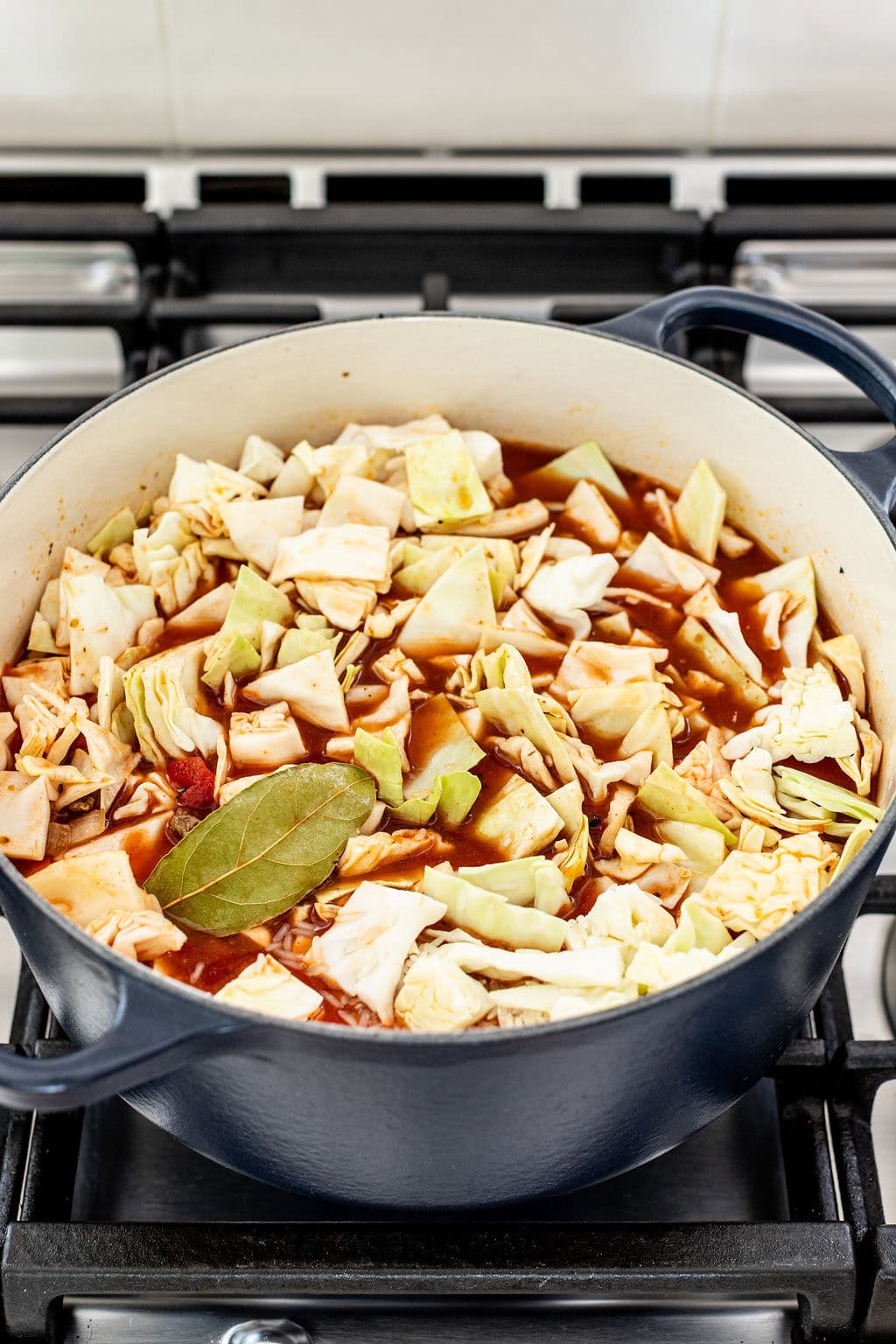 A large pot of soup with chopped cabbage, a bay leaf, and broth sits on a stove, simmering.