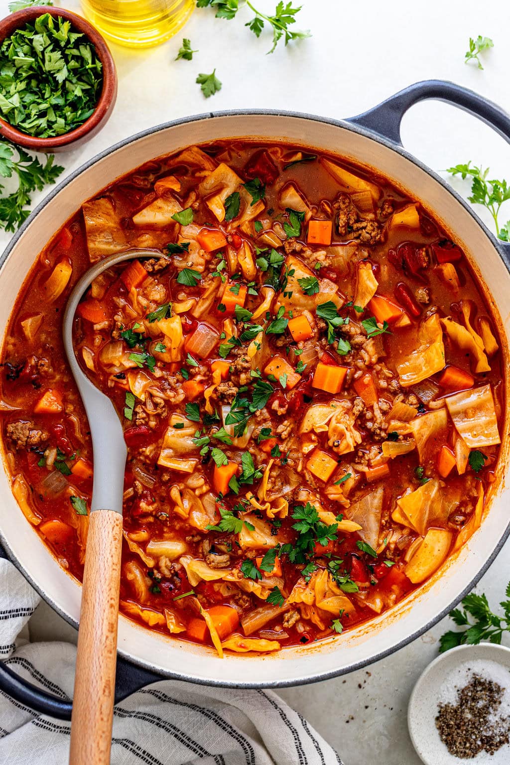 A pot of chunky vegetable soup with visible carrots, cabbage, tomatoes, and rice, garnished with chopped parsley. A ladle rests inside the pot; herbs and pepper sit nearby.