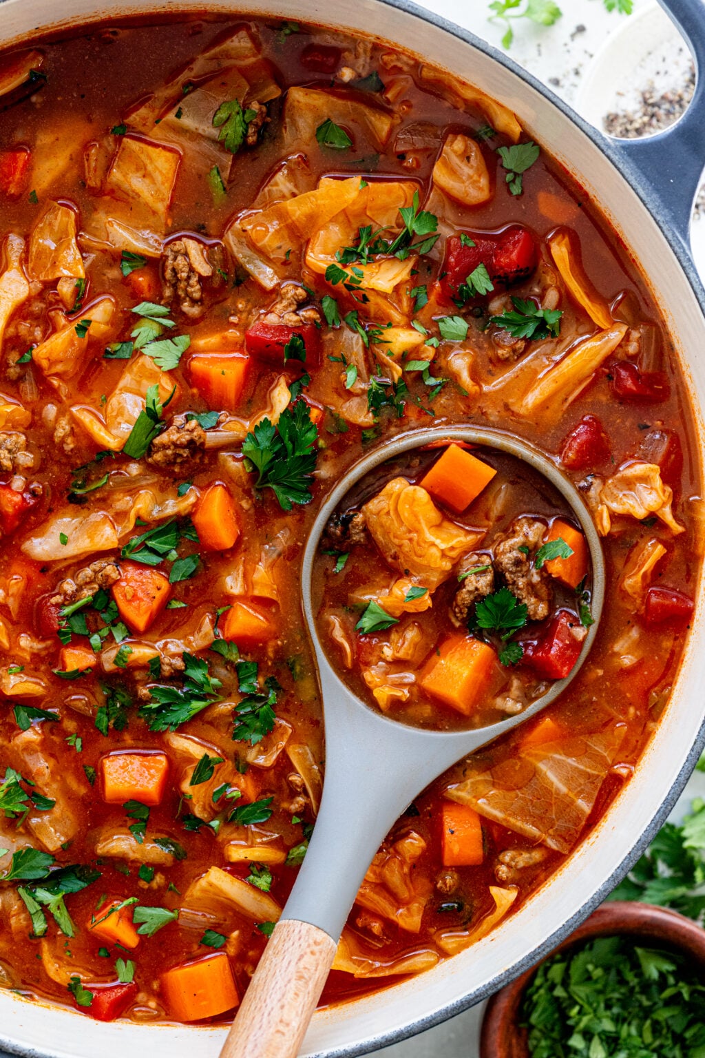 A pot of hearty soup with chunks of beef, carrots, cabbage, tomatoes, and fresh herbs, with a ladle lifting a serving.