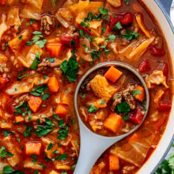 A pot of hearty soup with chunks of beef, carrots, cabbage, tomatoes, and fresh herbs, with a ladle lifting a serving.