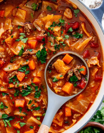 A pot of hearty soup with chunks of beef, carrots, cabbage, tomatoes, and fresh herbs, with a ladle lifting a serving.